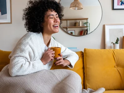 Frau mit Decke und Tasse in der Hand auf der Couch.