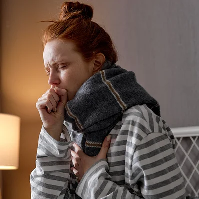 Frau mit Schal sitzt im Bett und hat Husten.