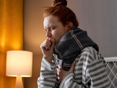 Woman wearing a scarf is sitting in bed and coughing.