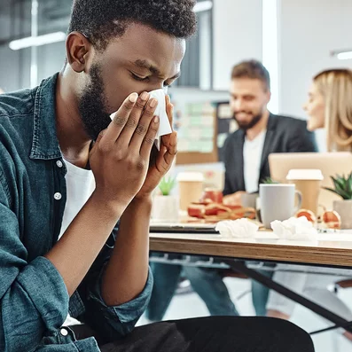 Mann mit Erkältung schnäuzt sich im Großraumbüro die Nase.