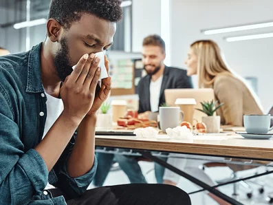 Mann mit Erkältung schnäuzt sich im Großraumbüro die Nase.