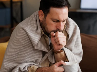 Husten verläuft bei einer Erkältung in verschiedenen Phasen Mann mit Erkältung und Husten sitzt mit Decke und Tee auf einer Couch.