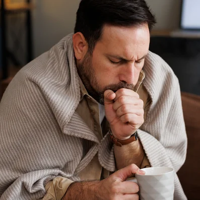 Mann mit Erkältung und Husten sitzt mit Decke und Tee auf einer Couch.